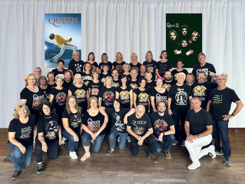 Der Chor Crescendo in schwarzen Queen-T-Shirts vor einem hellen Vorhang, links ein Plakat mit Freddie Mercury, rechts das Plakat zum Album „Queen II“
