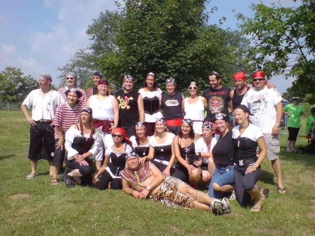 Gemischte Gruppe in Piratenkostümen mit Kopftüchern, Korsagen und Totenkopfmotiven auf einer Wiese, davor liegt ein Mitwirkender im Tigermuster-Kostüm