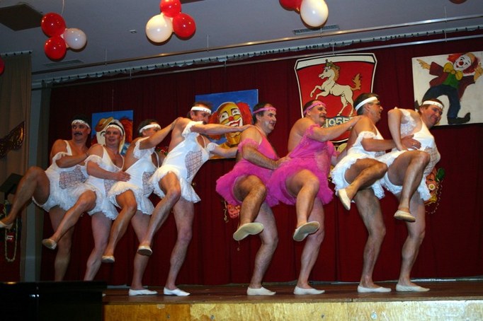 Das Männerballett in weißen und rosa Tüllkleidern mit Stirnbändern beim Tanz in einer Reihe auf der Bühne, ein Bein hochgeworfen