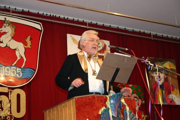 Ein Redner mit goldener Schärpe und Fliege am Rednerpult der Bürgerhausbühne, dahinter das Fastnachtswappen mit Schimmel und der Jahreszahl 1955