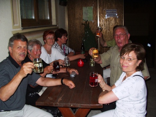 Sechs Reiseteilnehmer sitzen abends an einem Holztisch im Freien und heben ihre Gläser und Krüge in die Kamera