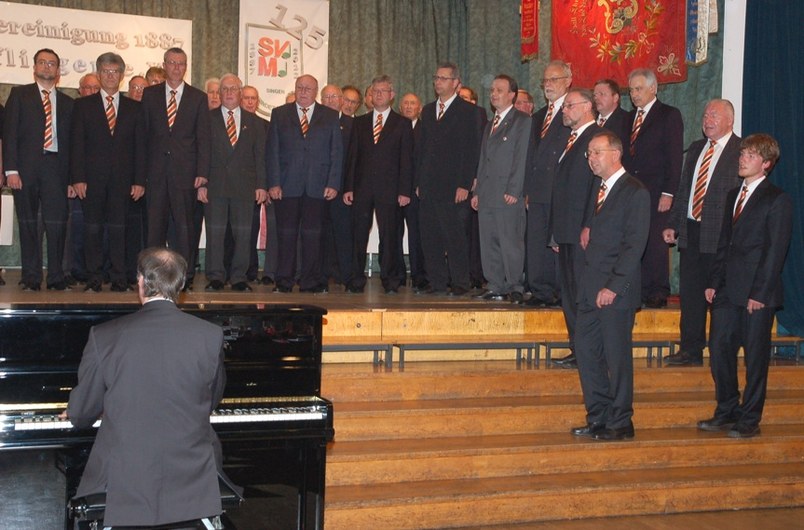 Der Männerchor in dunklen Anzügen mit gestreiften Krawatten auf der Bühne vor dem 125-Jahre-Schriftzug, vorn ein Pianist am Flügel