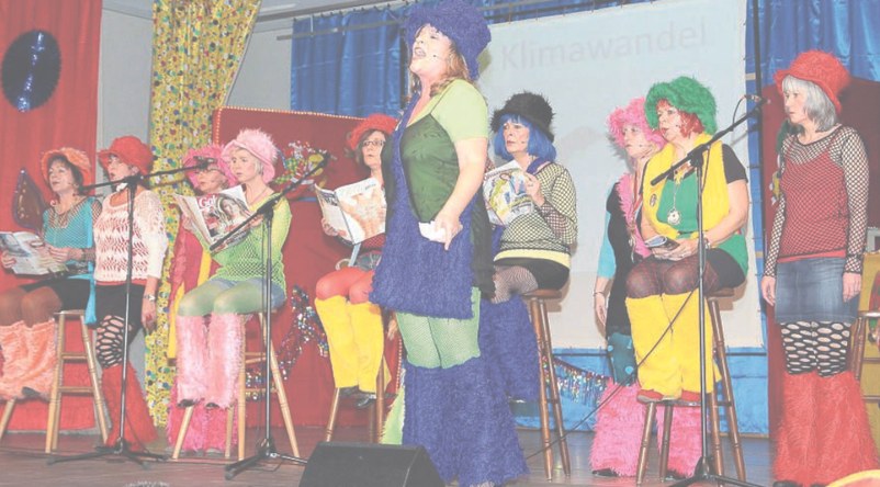 Die Moafährladies in bunten Kostümen mit Plüschhüten an Mikrofonen, Zeitschriften in den Händen, dahinter die Projektion „Klimawandel“