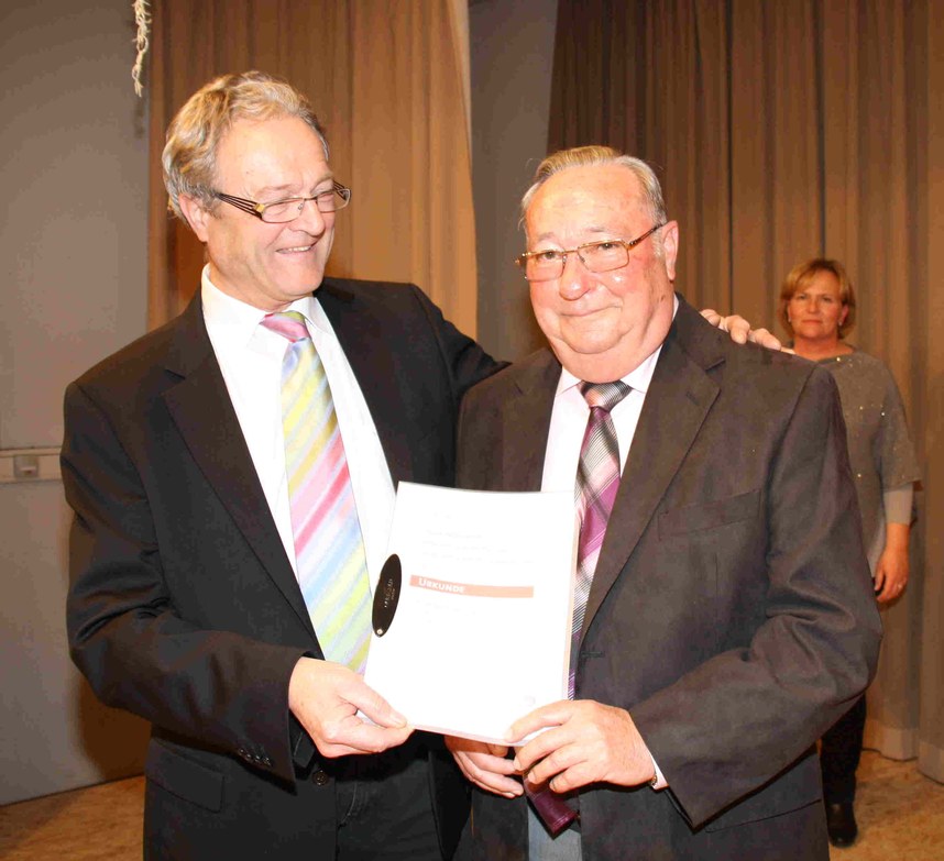 Ein Gratulant überreicht einem Geehrten lächelnd eine Urkunde und legt ihm dabei den Arm um die Schulter