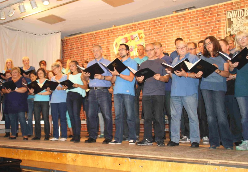 Sängerinnen und Sänger in Blautönen singen aus schwarzen Notenmappen auf der Bühne vor der Ziegelwand-Kulisse