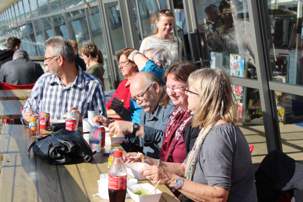 Mitglieder der Reisegruppe sitzen in der Sonne an langen Tischen hinter einer Glasfront und lassen sich Snacks und Getränke schmecken