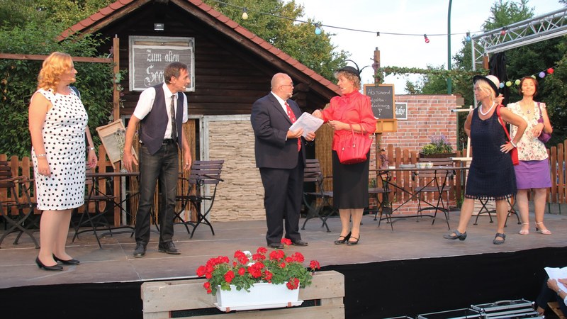 Sechs Darsteller vor der Kulisse eines Seelokals auf der Freilichtbühne, in der Mitte der Bürgermeister im Anzug mit Papieren neben einer Dame in roter Bluse