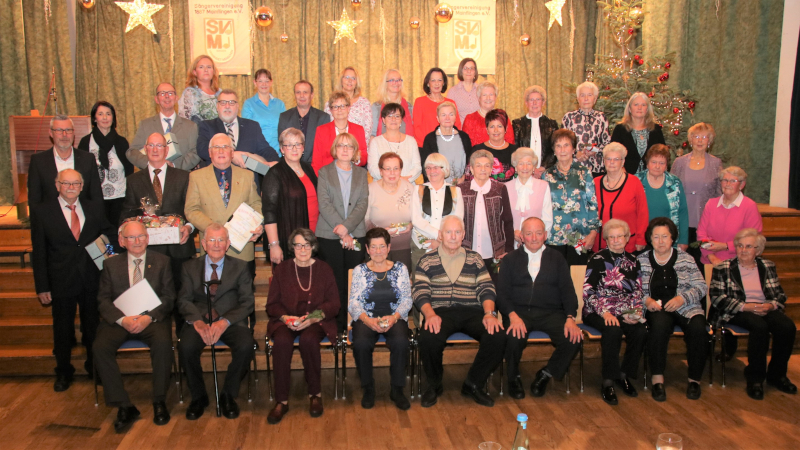 Gruppenbild der Geehrten im weihnachtlich geschmückten Saal mit Sternen und Christbaum, vorn eine sitzende Reihe, dahinter zwei Reihen stehend
