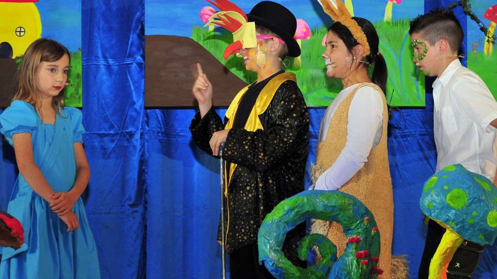 Szene aus dem Kindermusical: Alice im blauen Kleid trifft vor der bunten Wunderland-Kulisse auf Kinder in Vogel-, Hasen- und Eidechsenkostümen