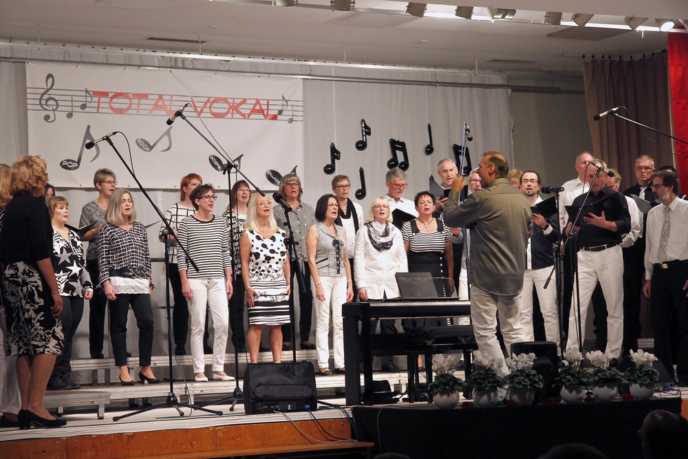Ein großer gemischter Chor in schwarz-weißer Kleidung singt vor dem Banner „Total Vokal“ auf der Bürgerhausbühne, vorne der Dirigent am Keyboard