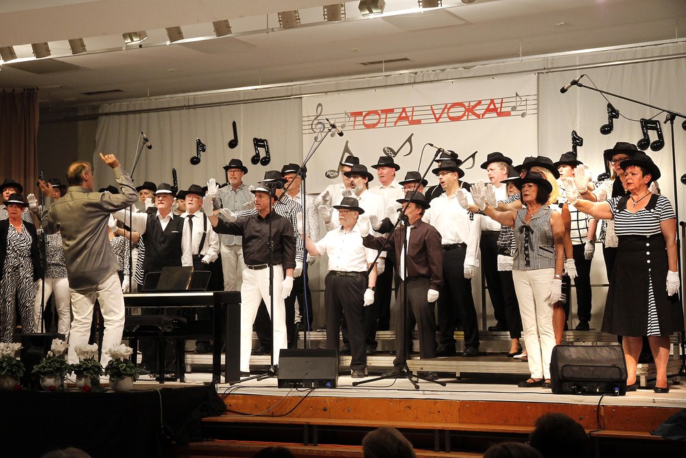 Der Dirigent mit erhobener Hand vor dem Chor, der mit schwarzen Hüten und weißen Handschuhen unter dem Banner „Total Vokal“ eine Schau-Nummer singt