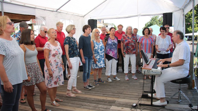 Ein Chor singt unter dem weißen Zeltdach auf der Holzterrasse, rechts begleitet ein Pianist am E-Piano