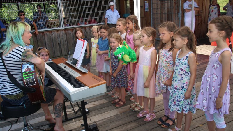 Der Kinderchor singt auf der Holzterrasse, ein Mädchen hält eine grüne Krokodil-Handpuppe, links begleitet die Chorleiterin am E-Piano