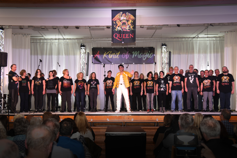 Ein Solist im gelben Jackett und weißer Hose im Freddie-Mercury-Look vor dem Chor auf der Bühne