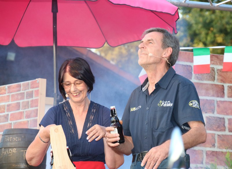 Eine Darstellerin öffnet lachend eine Flasche, neben ihr ein Darsteller im dunklen Hemd